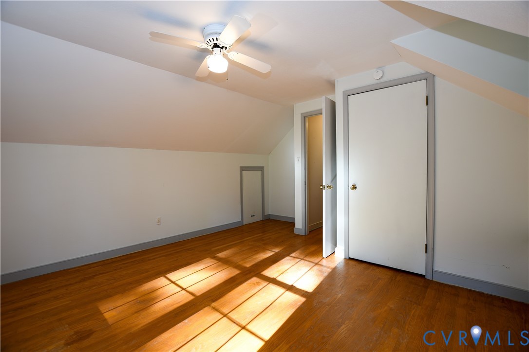 Room featuring wood-finish flooring, vaulted ceilings with a center-mounted ceiling fan, built-in wall storage, and interior door access