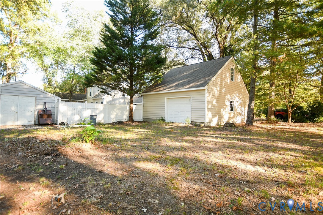 Detached multi-level garage with lap siding, asphalt shingle roof, and white sectional garage door, accompanied by an auxiliary storage structure and perimeter white vinyl fencing
