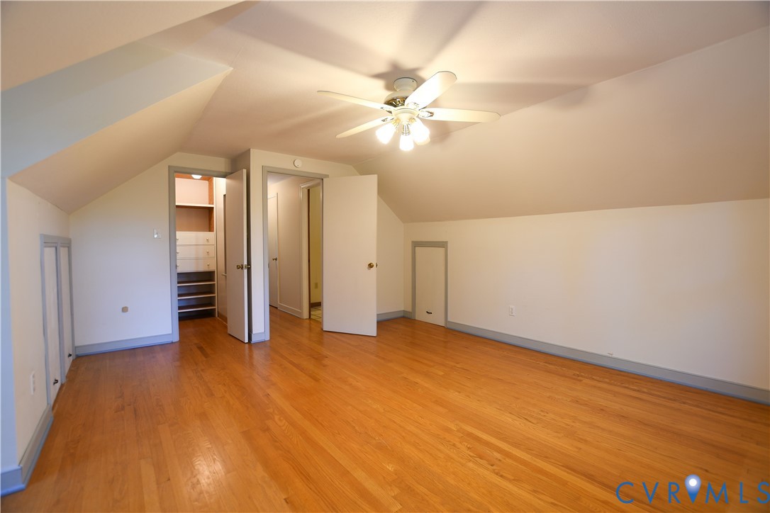 Room featuring wood-finish flooring, vaulted ceilings, a ceiling fan with integrated lighting, built-in wall storage, and interior access doors