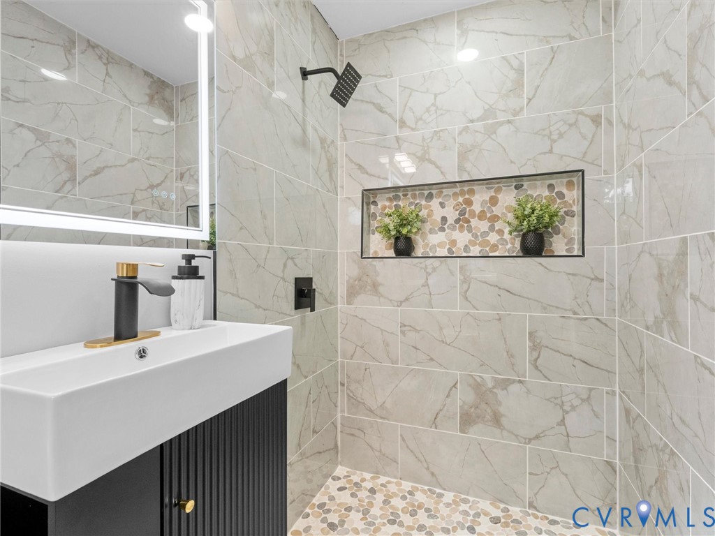 Bathroom vanity featuring a fluted dark cabinet, white rectangular basin, and matte black faucet, accompanied by a shower enclosure with large-format stone-finish tile walls, pebble-stone flooring, and a recessed tiled niche