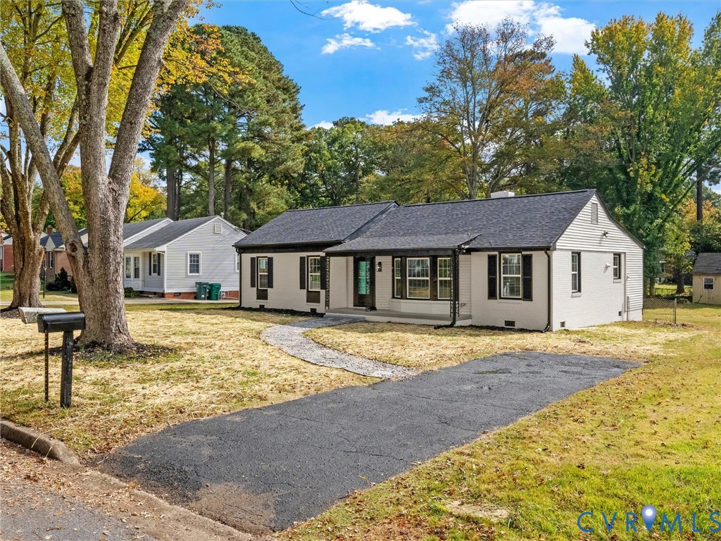 Single-story residence featuring light-colored exterior siding, a dark-toned shingled roof, contrasting window shutters, a stone walkway, and an asphalt driveway Single-story residence featuring light-colored exterior siding, a dark-toned shingled roof, contrasting window shutters, a stone walkway, and an asphalt driveway