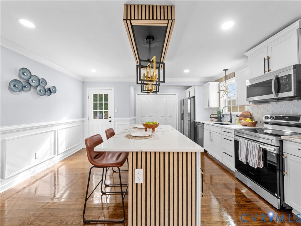 Kitchen featuring a center island with slatted wood detailing and a stone-style countertop, custom vertical-slat pendant lighting, shaker-style cabinetry, stainless steel appliances, and wainscoting