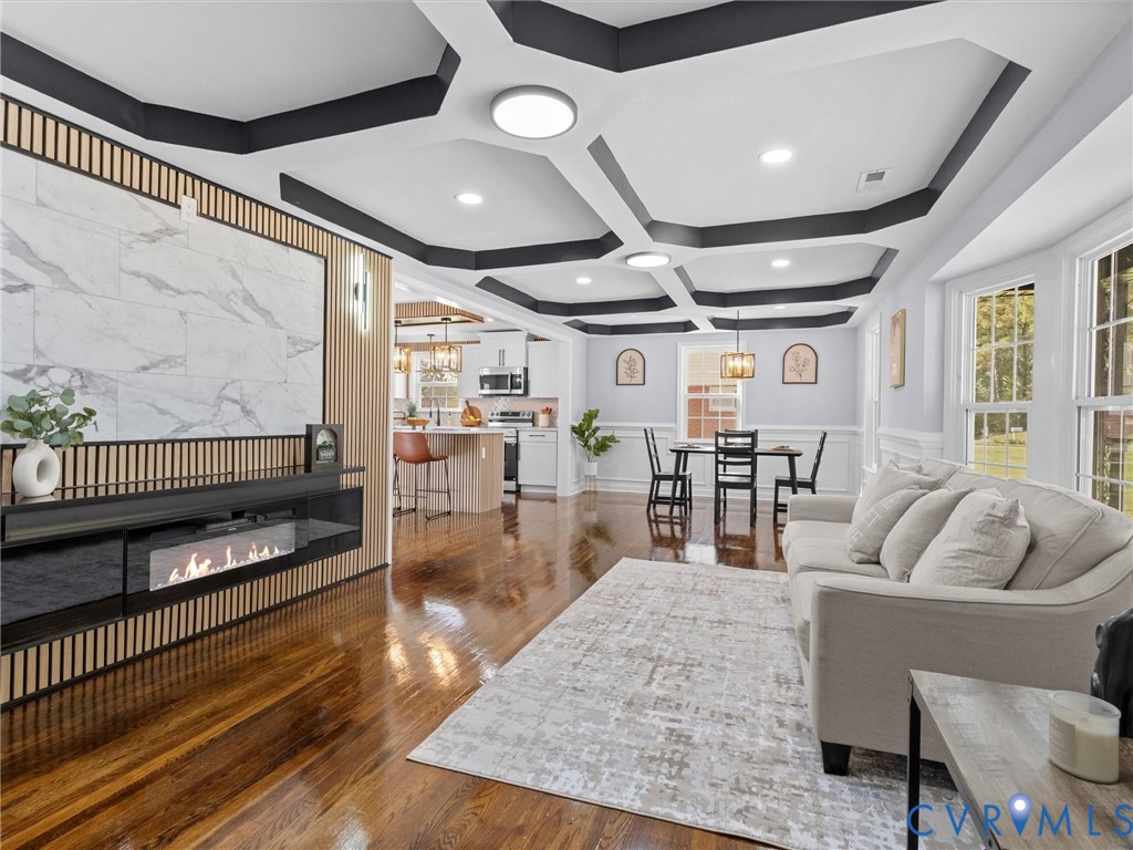 Coffered ceiling with dark accent trim, wood-finish flooring, custom slatted wood wall feature, integrated electric fireplace with stone-look surround, and recessed lighting Coffered ceiling with dark accent trim, wood-finish flooring, custom slatted wood wall feature, integrated electric fireplace with stone-look surround, and recessed lighting
