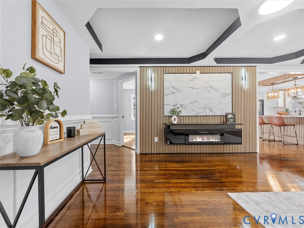 Wood-finish flooring, tray ceilings with contrasting trim, and a floor-to-ceiling vertical slat accent wall featuring a stone-style panel and built-in electric fireplace Wood-finish flooring, tray ceilings with contrasting trim, and a floor-to-ceiling vertical slat accent wall featuring a stone-style panel and built-in electric fireplace