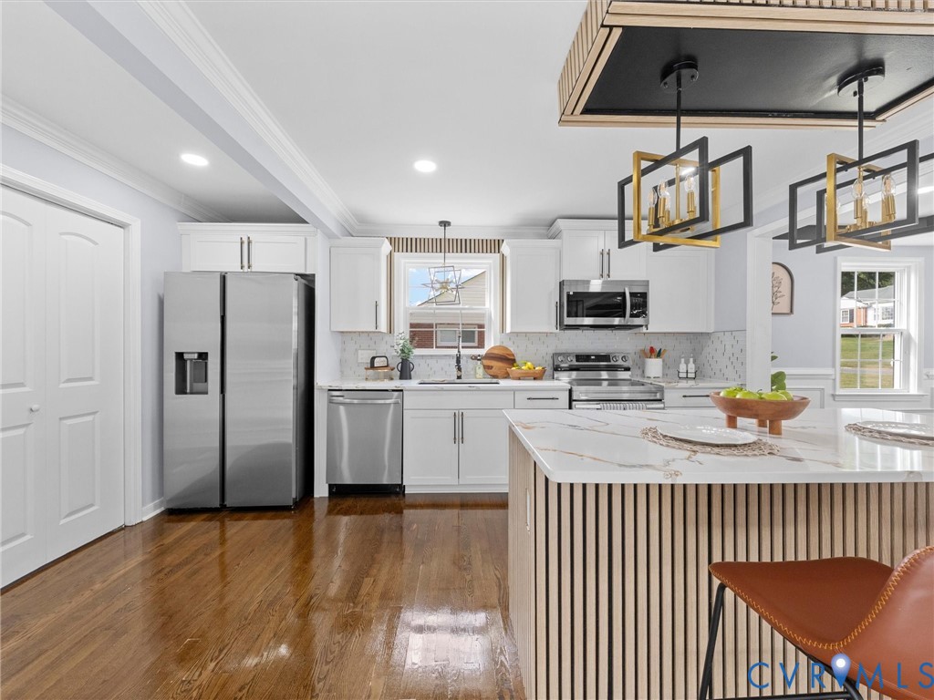 Kitchen featuring white shaker-style cabinetry, stainless steel appliances, wood-finish flooring, a kitchen island with a slatted wood-finish base, and geometric pendant lighting