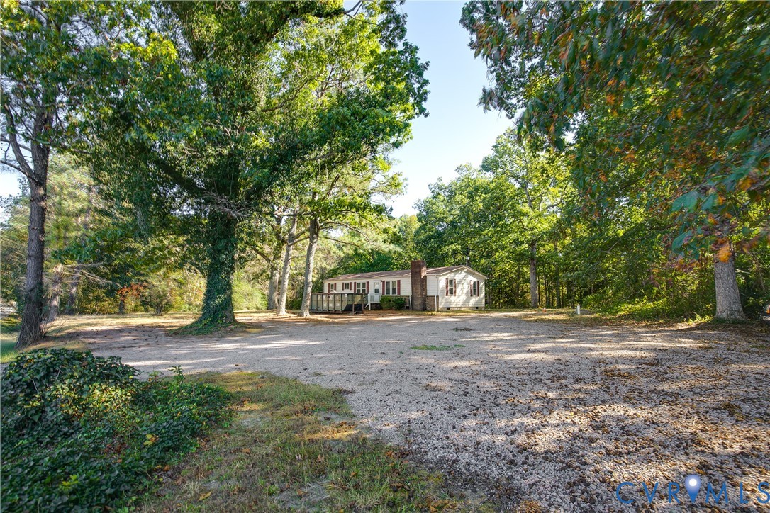 Single-story exterior featuring light-toned siding, a brick chimney, an elevated wooden deck, and an expansive gravel driveway situated within a mature wooded lot Single-story exterior featuring light-toned siding, a brick chimney, an elevated wooden deck, and an expansive gravel driveway situated within a mature wooded lot