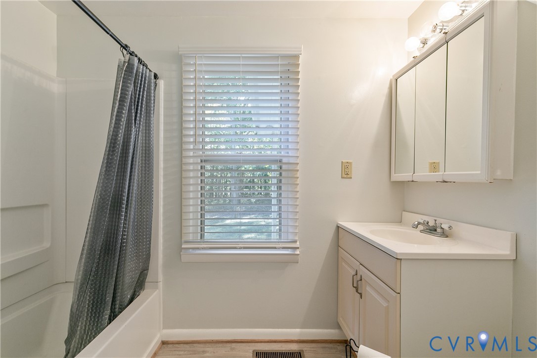 Bathroom featuring a white vanity cabinet with an integrated sink, a three-door mirrored medicine cabinet, a light-toned wood-finish floor, and a white tub-shower combination with horizontal blinds