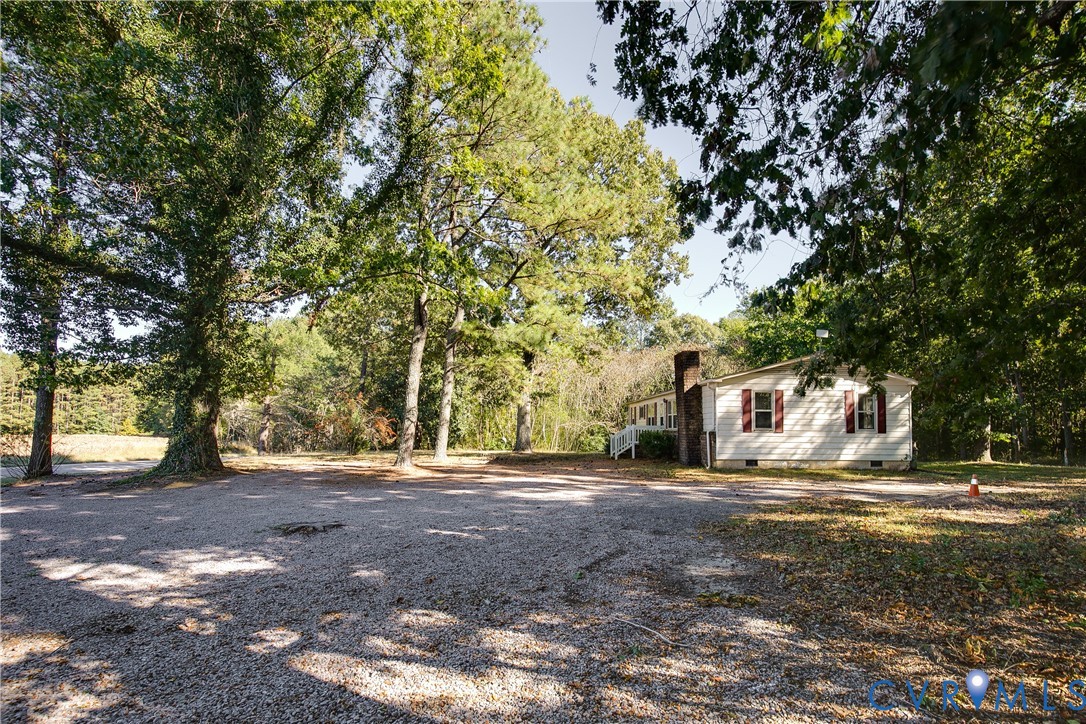 Exterior featuring light-colored horizontal siding, red shutters, a brick chimney, and a gravel lot surrounded by mature trees Exterior featuring light-colored horizontal siding, red shutters, a brick chimney, and a gravel lot surrounded by mature trees
