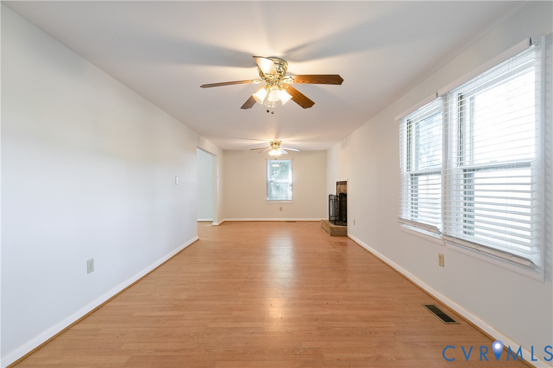 Living area featuring wood-finish flooring, two ceiling fans, large windows with horizontal blinds, and a fireplace with a stone-finish hearth Living area featuring wood-finish flooring, two ceiling fans, large windows with horizontal blinds, and a fireplace with a stone-finish hearth