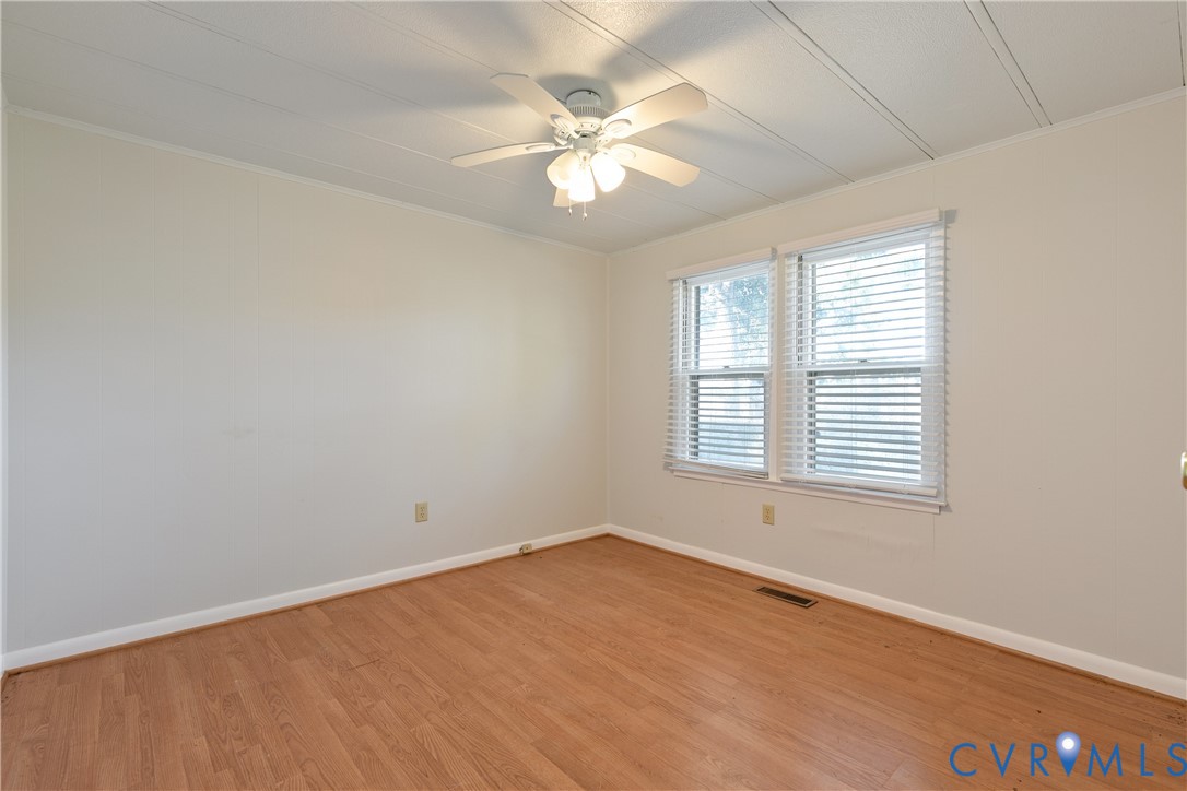Neutral-toned interior space featuring wood-finish flooring, white wall paneling, a multi-blade ceiling fan with integrated lighting, and dual windows equipped with horizontal blinds