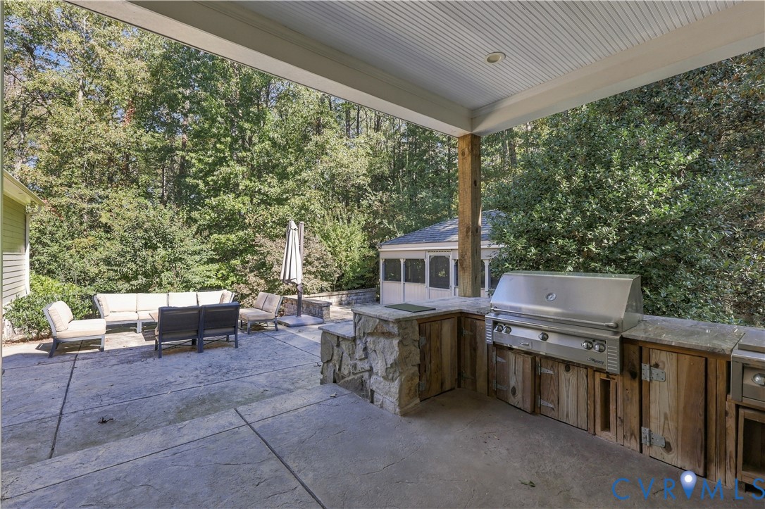 Covered patio featuring a built-in outdoor kitchen with stone masonry, wood-finish cabinetry, a stainless steel grill, and a concrete paver surface