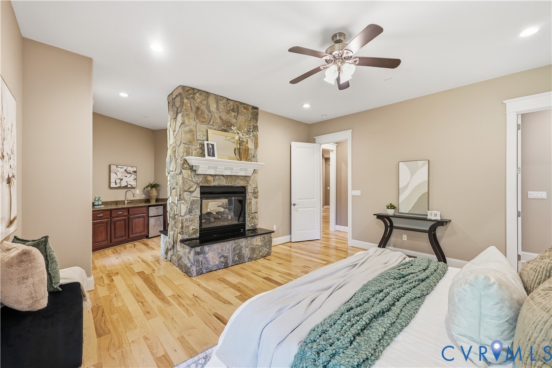 Stone-faced double-sided fireplace with a raised hearth, wood-finish flooring, recessed lighting, ceiling fan, and built-in wet bar featuring dark wood cabinetry and a sink