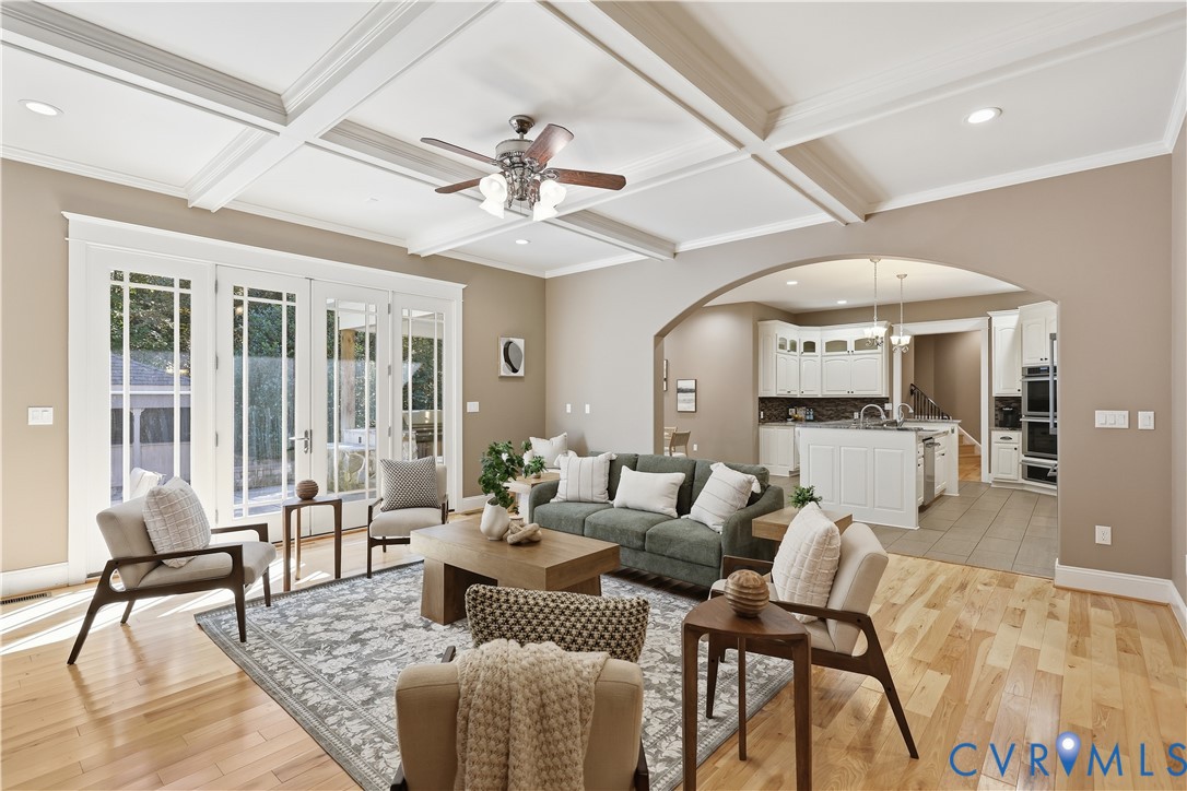 Coffered ceilings with recessed lighting, wood-finish flooring, multi-pane French doors, and an arched entryway leading to a kitchen with white cabinetry and a central island