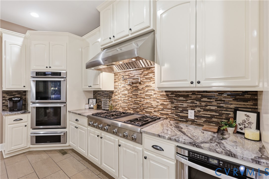 Kitchen featuring white cabinetry, stone-surface countertops, horizontal tile backsplash, stainless steel double wall ovens, and a professional-style gas range with a stainless steel vent hood