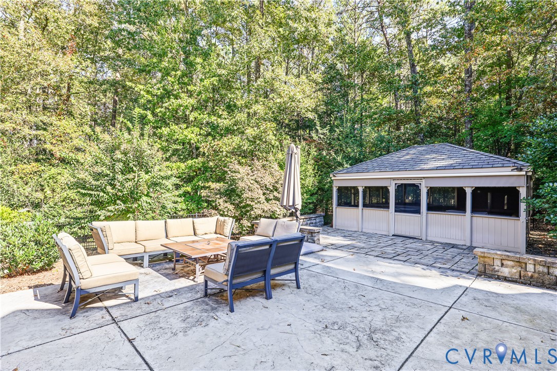 Stamped concrete patio featuring a detached screened pavilion, stacked stone retaining wall, and mature wooded perimeter