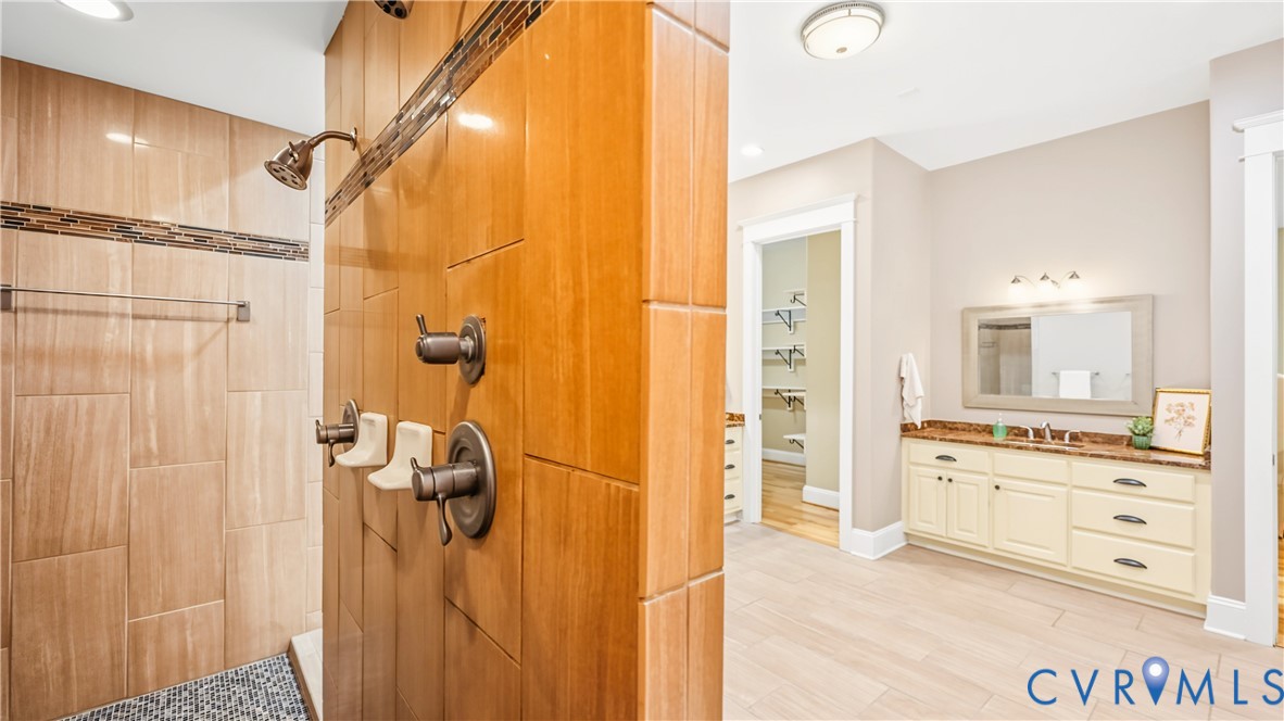 Walk-in shower featuring wood-finish tile walls, stone mosaic floor inlay, and bronze-toned hardware, paired with a dual-sink vanity topped with stone-finish surfacing