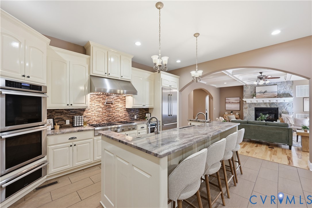 Kitchen featuring off-white cabinetry, stone-finish countertops, stainless steel appliances, a textured tile backsplash, and a central island with a prep sink