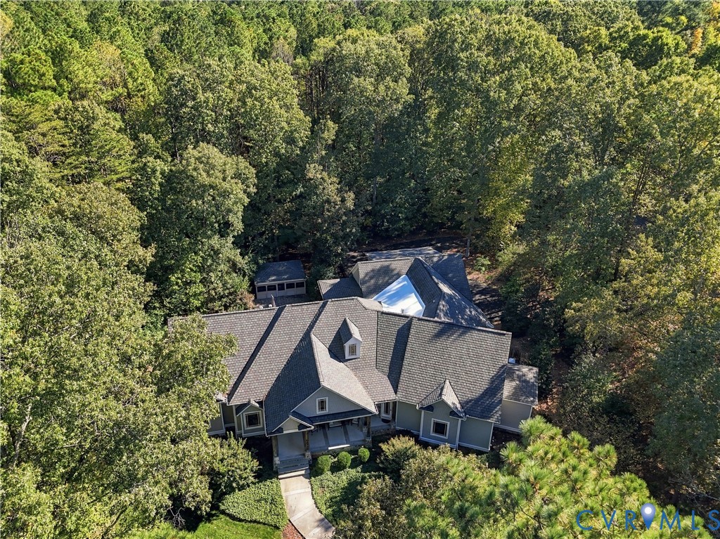 Aerial perspective of a multi-gabled residence featuring shingle roofing, exterior siding, a concrete walkway, and a detached structure, all situated on a heavily wooded lot