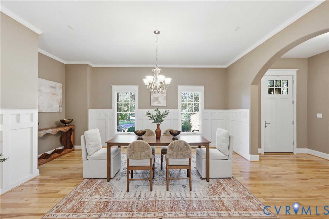 Dining area featuring wood-finish flooring, white wainscoting, a multi-arm chandelier, dual windows with decorative casing, and an arched entryway