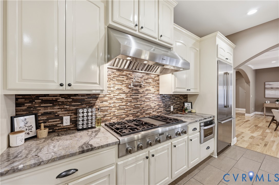 Kitchen featuring white cabinetry, stone countertops, multi-toned tile backsplash, stainless steel gas range with overhead vent hood, and stainless steel refrigerator