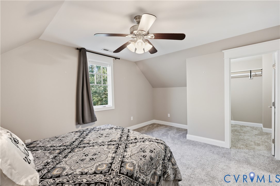 Upstairs Bedroom. Neutral wall palette featuring an angled ceiling, wall-to-wall carpeting, a multi-blade ceiling fan with integrated lighting, a multi-pane window, and a walk-in closet with built-in shelving