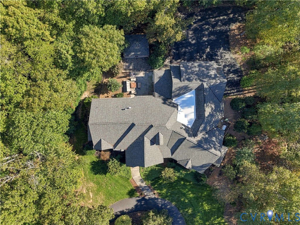Aerial perspective of a multi-gabled architectural roofline, paved driveway, concrete hardscaping, and mature woodland canopy on a private lot