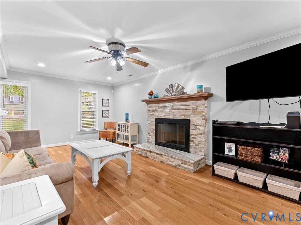 Living space featuring wood-finish flooring, a stacked-stone fireplace with a wooden mantel, a stone hearth, and a ceiling fan with integrated lighting