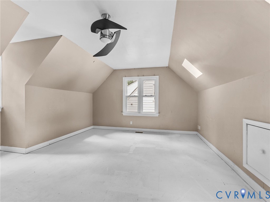 Vaulted ceiling room featuring a skylight, multi-pane window, modern matte black ceiling fan, and access door to built-in attic storage