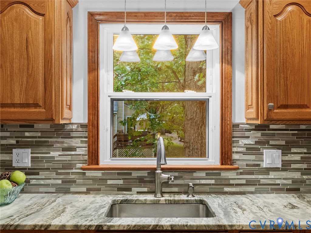 Kitchen features include wood-finish cabinetry, a natural stone countertop, an undermount stainless steel sink with a high-arch faucet, a multi-tonal linear tile backsplash, and triple pendant lighting