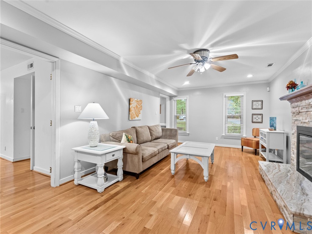 Wood-finish flooring, stone-faced fireplace with a natural stone hearth, crown molding, and ceiling fan with integrated lighting