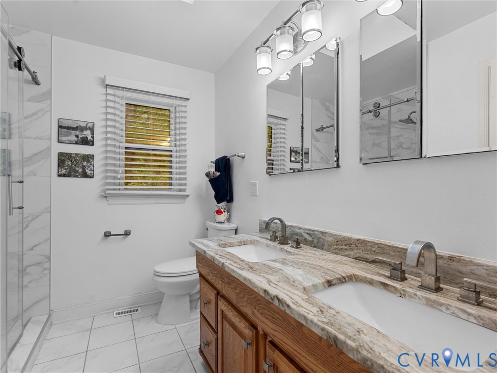 Double vanity featuring a stone countertop, dual undermount sinks with metallic faucets, wood-finish cabinetry, and a walk-in shower with floor-to-ceiling tile surround