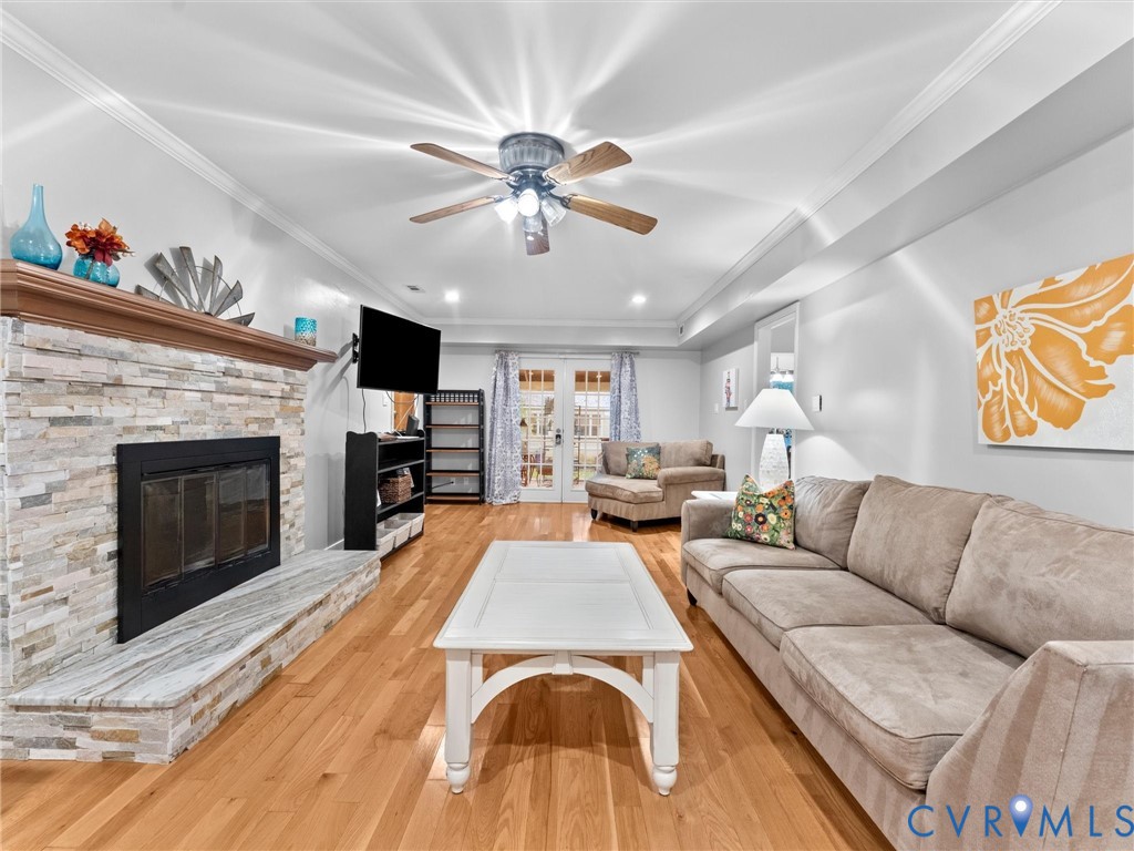 Stacked stone fireplace with a stone hearth, wood-finish flooring, ceiling fan with integrated lighting, crown molding, and glass-paneled exterior doors