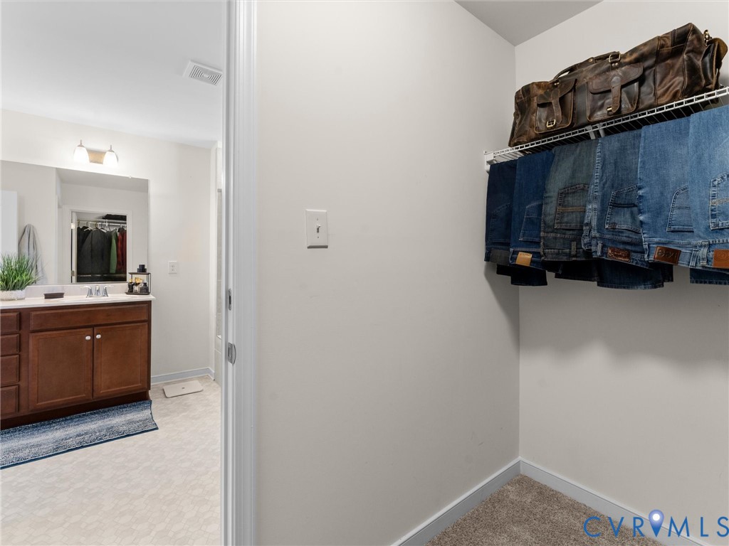 Walk-in closet featuring wire shelving, wall-to-wall carpeting, and an entryway into a bathroom equipped with a wood-finish vanity, solid-surface countertop, and wall-mounted vanity lighting