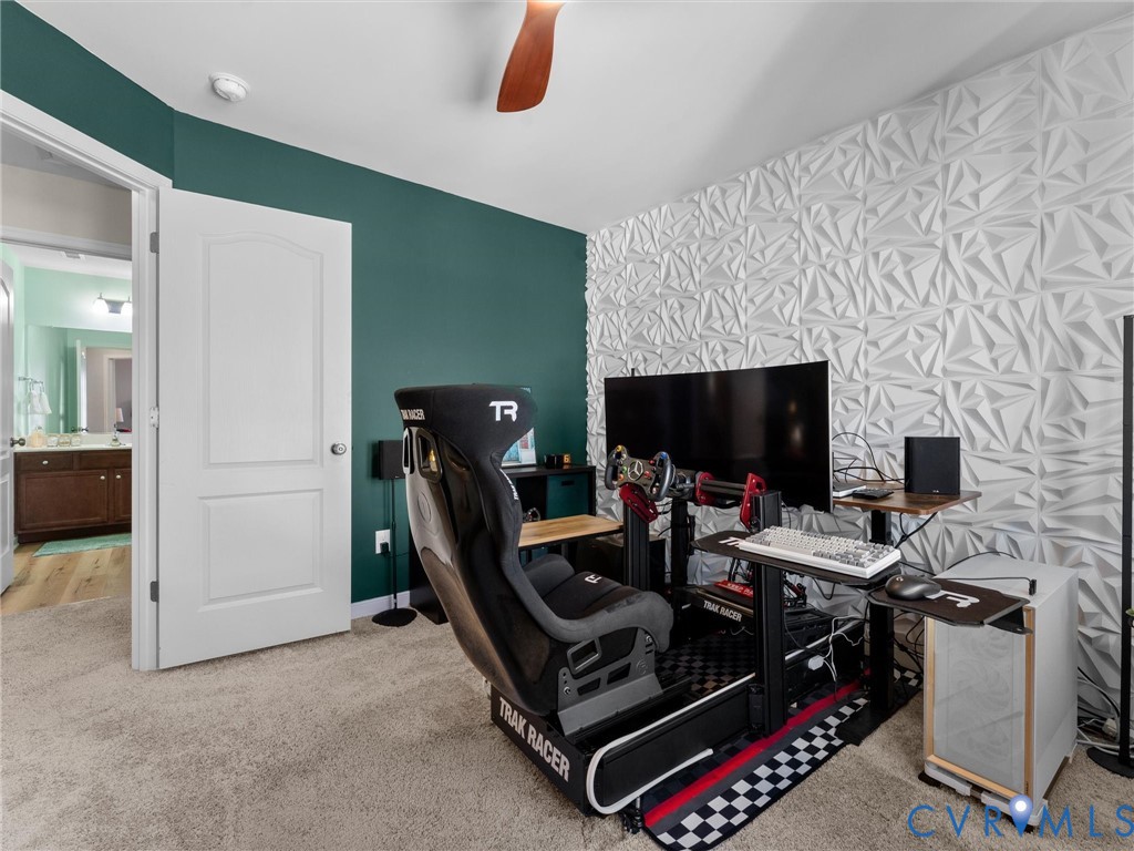 Room featuring an accent wall with geometric three-dimensional paneling, deep green painted wall, neutral carpeting, and a ceiling fan with wood-finish blades
