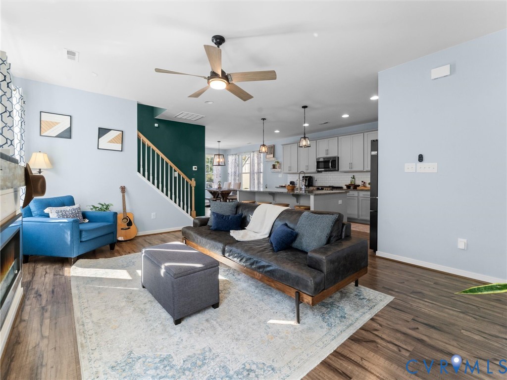 Open-concept interior featuring wood-finish flooring, a kitchen island, pendant lighting, a ceiling fan, and a staircase with a wooden handrail