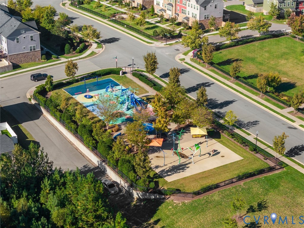 Community recreation area featuring a paved playground, water play elements, shade structures, and mature trees enclosed by perimeter fencing