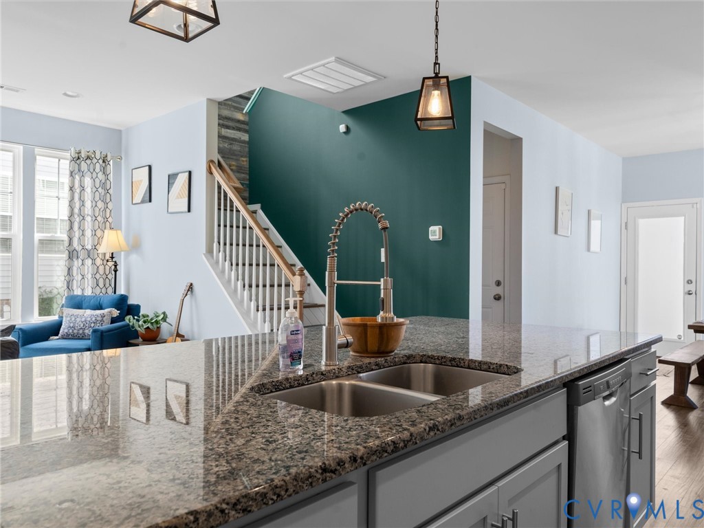 Kitchen island with stone-surface countertop, stainless steel dual-basin undermount sink, gooseneck faucet, gray cabinetry, and wooden staircase with white balusters