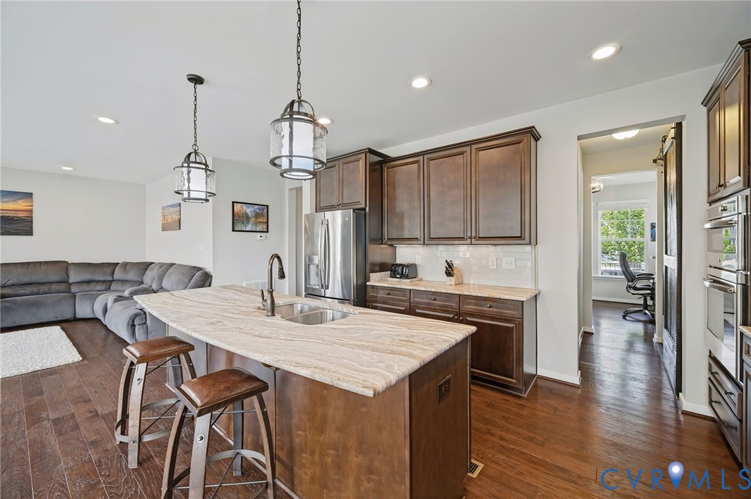 Kitchen featuring stained wood cabinetry, a neutral-toned stone-finish countertop island with an undermount sink, recessed lighting, pendant fixtures, and built-in stainless steel wall ovens