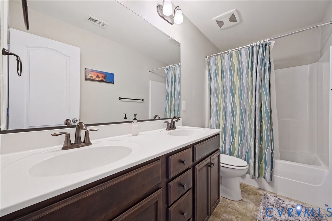Bathroom featuring a dual-sink vanity with dark wood cabinetry, solid-surface countertop, bronze-tone fixtures, full-width wall mirror, and a tub-shower combination