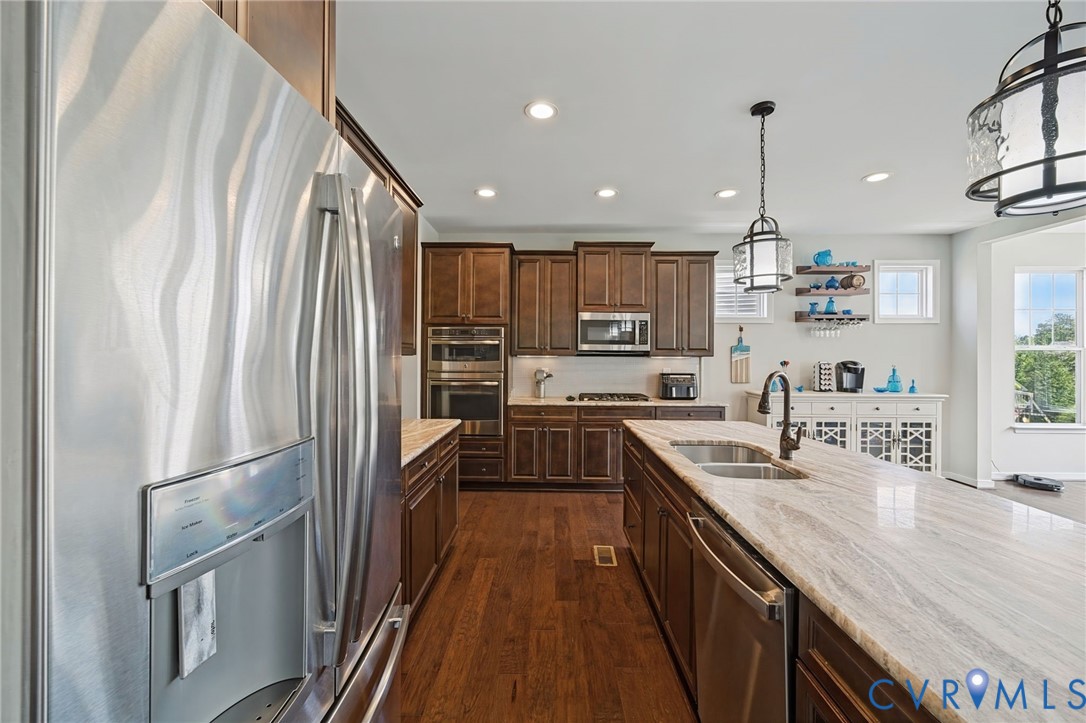 Kitchen featuring stained wood cabinetry, stone countertops, stainless steel appliances, a center island with an integrated sink, and wood-finish flooring