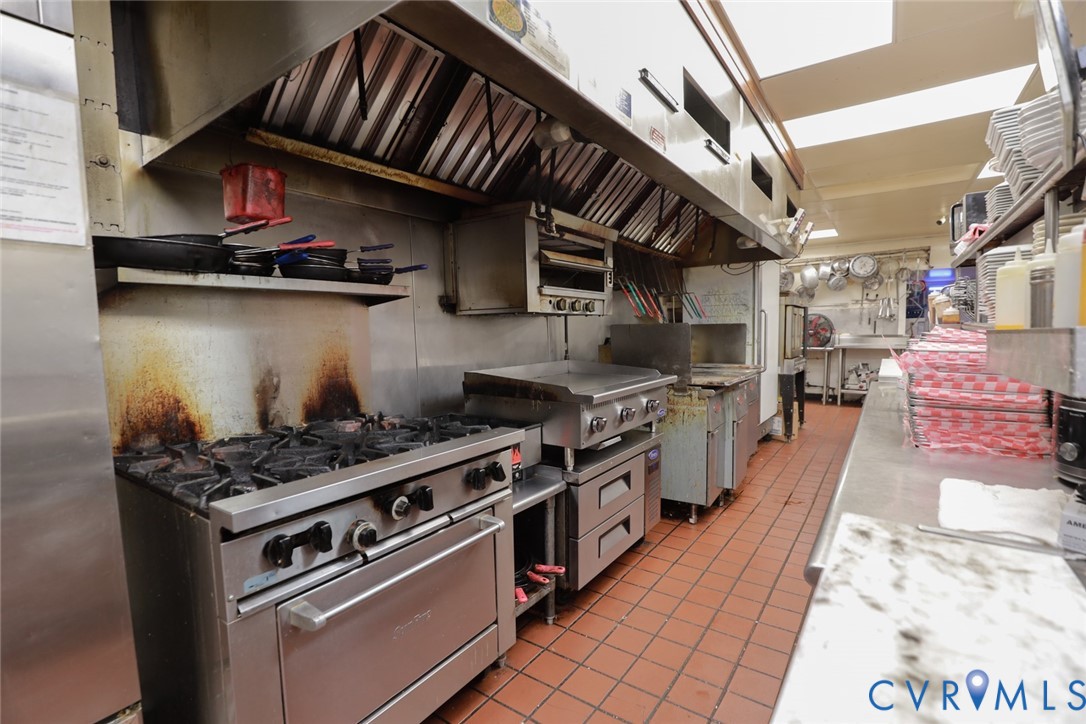 Commercial kitchen featuring a stainless steel ventilation hood, industrial-grade range with a built-in oven, flat-top grill, stainless steel prep surfaces, and tile flooring