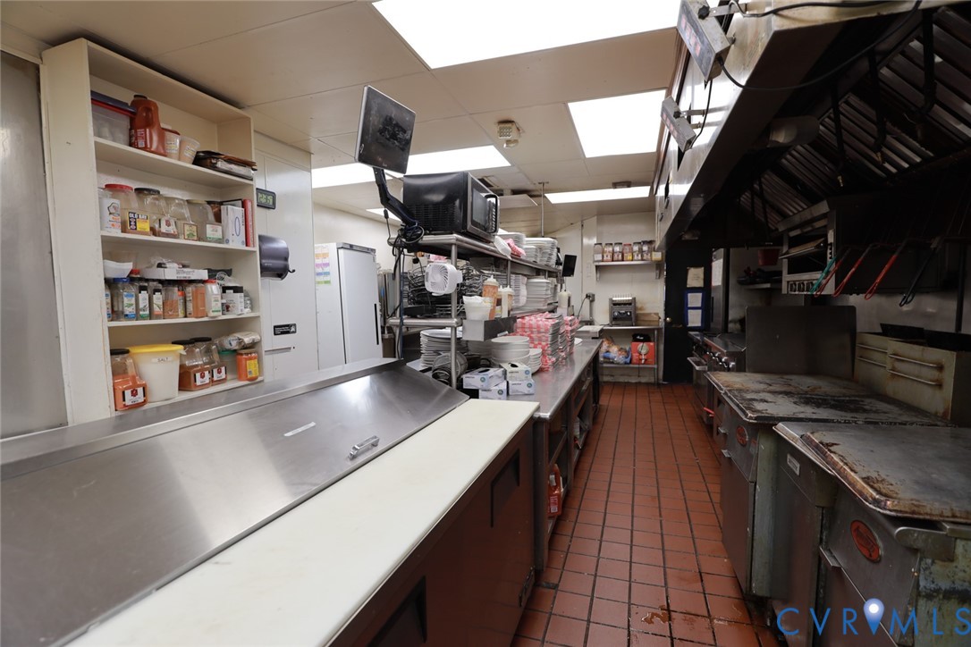 Commercial kitchen featuring stainless steel food preparation stations, built-in shelving, high-capacity ventilation hood, professional-grade range, and tile flooring