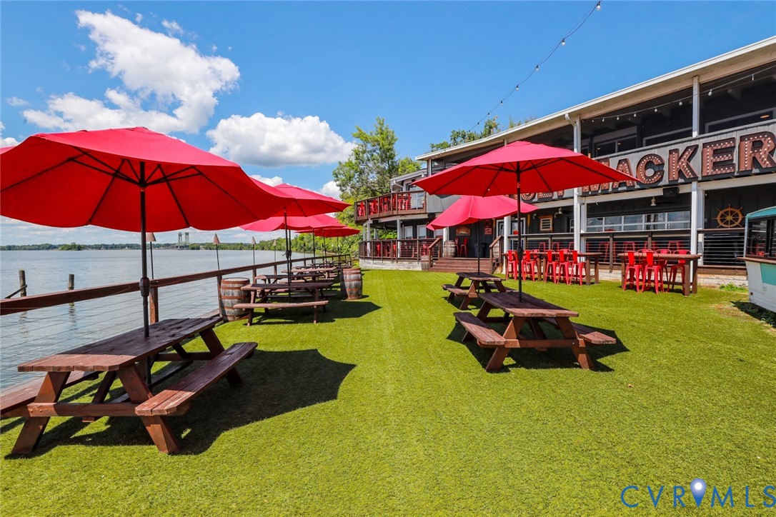 Waterfront outdoor space featuring an expansive artificial turf lawn, a perimeter wood railing, and an elevated building exterior with integrated string lighting