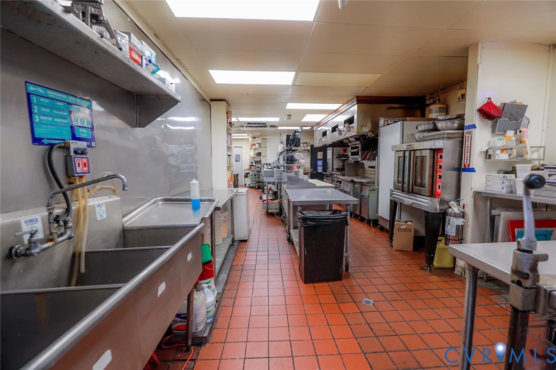 Commercial kitchen featuring stainless steel wall-mounted shelving, triple-basin metal sink, industrial-grade tile flooring, and integrated stainless steel convection ovens