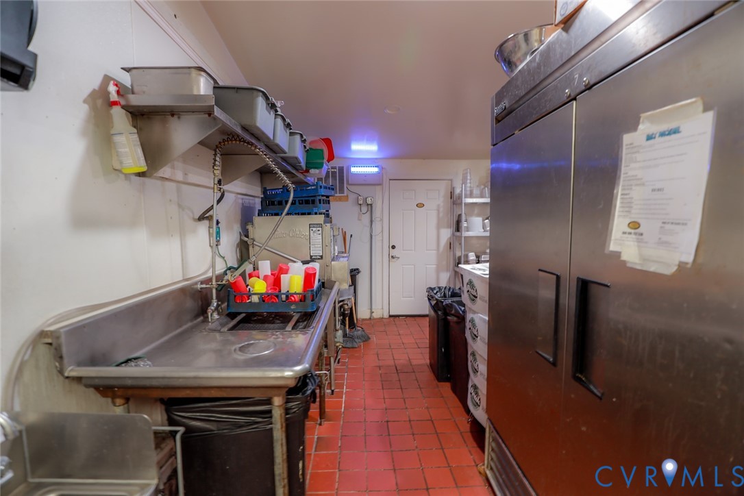 Commercial kitchen prep area featuring stainless steel dual-basin sink, overhead shelving, industrial-grade stainless steel refrigeration unit, and durable quarry tile flooring