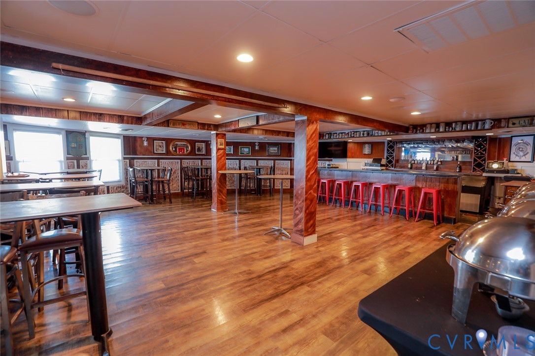 Spacious interior featuring wood-finish flooring, built-in bar with mirror backing, recessed ceiling lighting, and decorative wood-wrapped support pillars