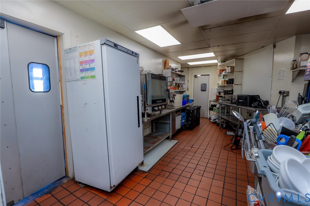 Commercial kitchen workspace featuring durable tile flooring, recessed panel lighting, stainless steel work surfaces, wall-mounted shelving, and a full-height refrigerator