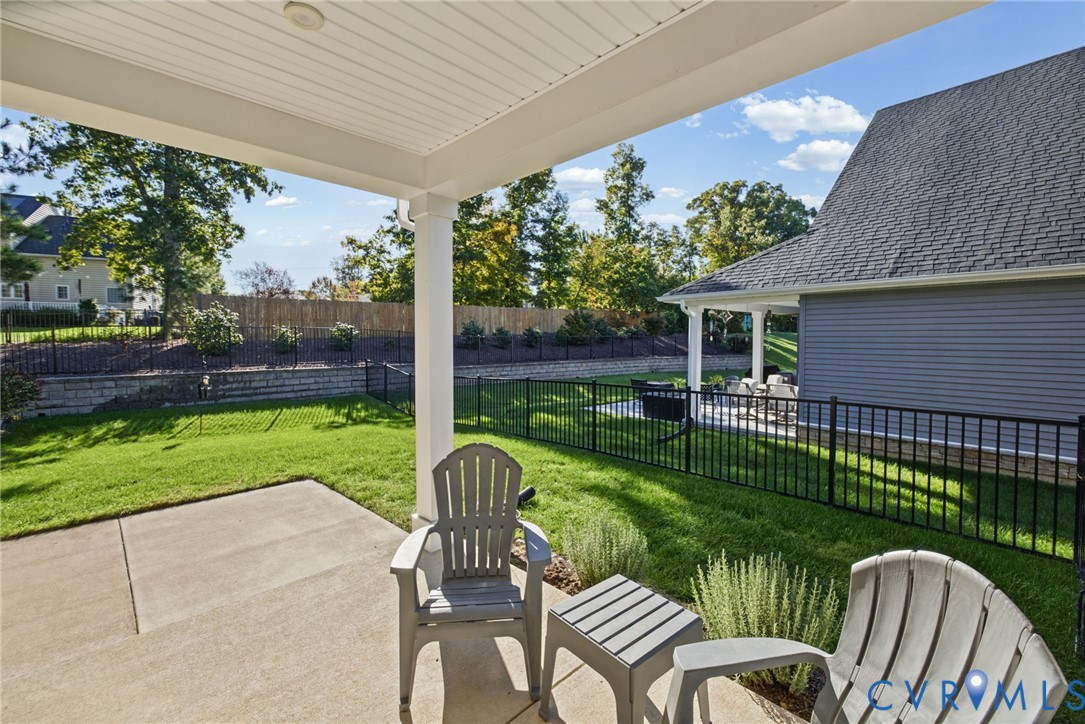 Covered concrete patio with a beadboard ceiling, manicured lawn, black metal perimeter fencing, and stone retaining wall
