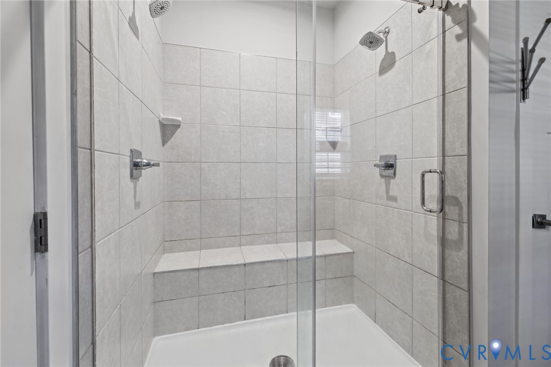 Walk-in shower featuring light-toned tile surround, built-in tiled bench, dual overhead showerheads, and upgraded frameless glass door.