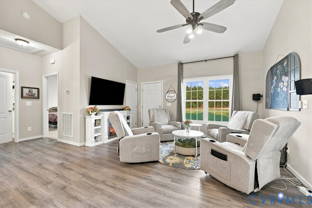 Living area featuring vaulted ceilings, wood-finish flooring, a built-in media console with an integrated fireplace, and a ceiling fan with multi-light fixture Living area featuring vaulted ceilings, wood-finish flooring, a built-in media console with an integrated fireplace, and a ceiling fan with multi-light fixture